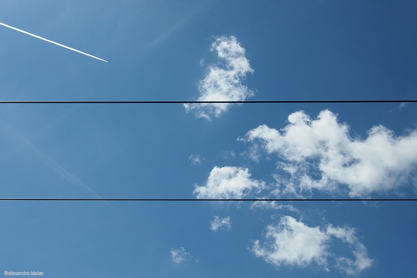 Blick auf blauen Himmel mit weißen Wolken, Kondensstreifen und zwei Stromleitungen, die horizontal verlaufen.